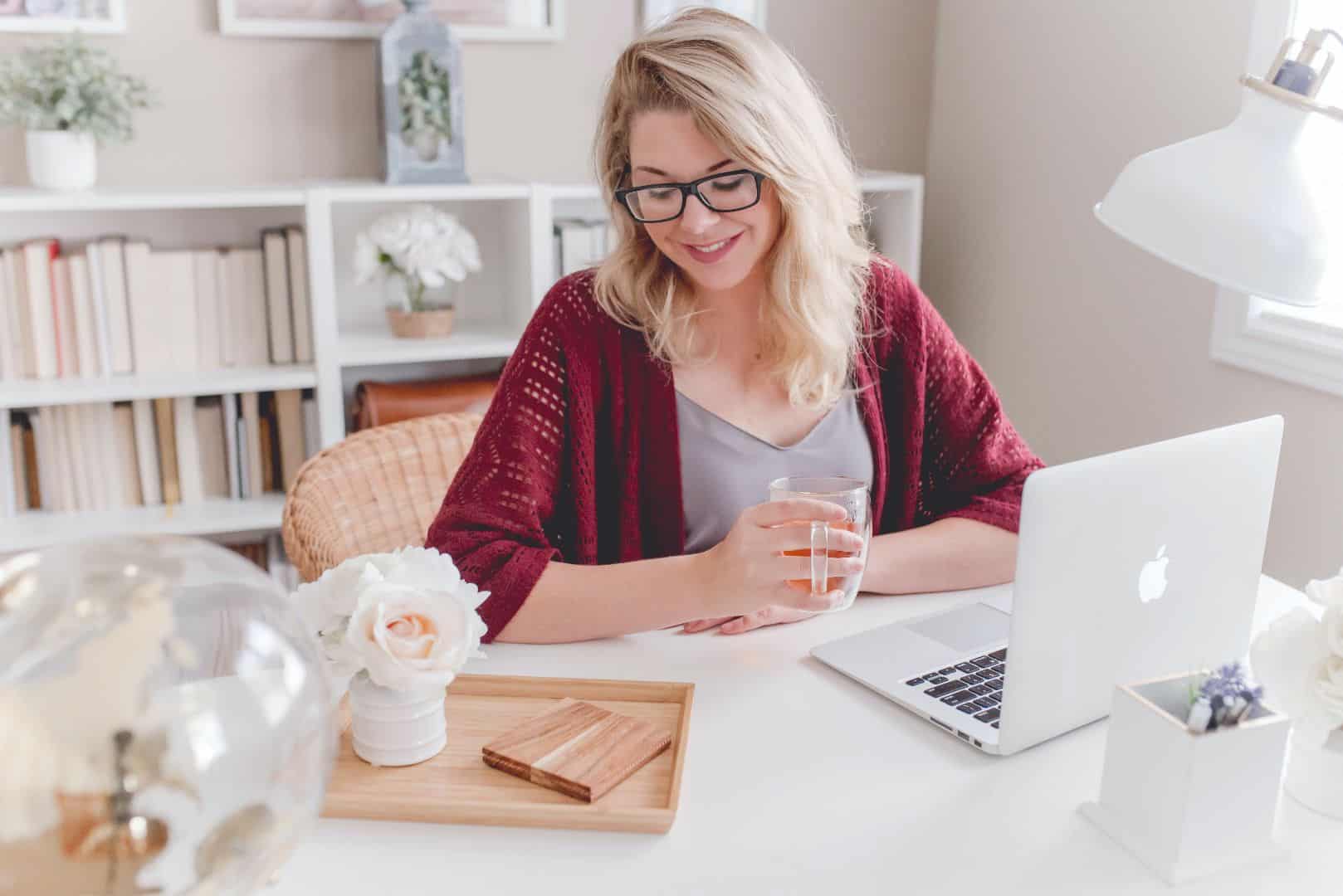 Relaxed woman working on a laptop at home, holding a glass of water, surrounded by decorative plants and flowers, in a bright and modern workspace with natural light.