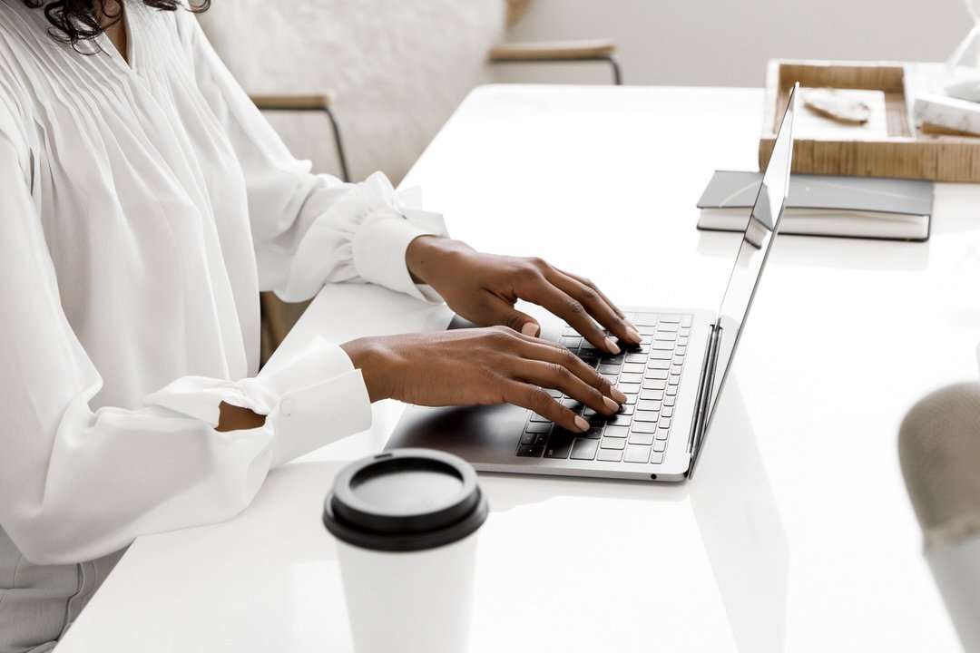 A woman engaged in typing on her laptop at a desk, surrounded by a few personal items and a bright atmosphere.