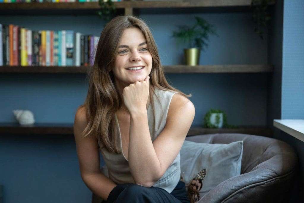 Professional woman smiling in an office or co-working space with a bookshelf and plants in the background.
