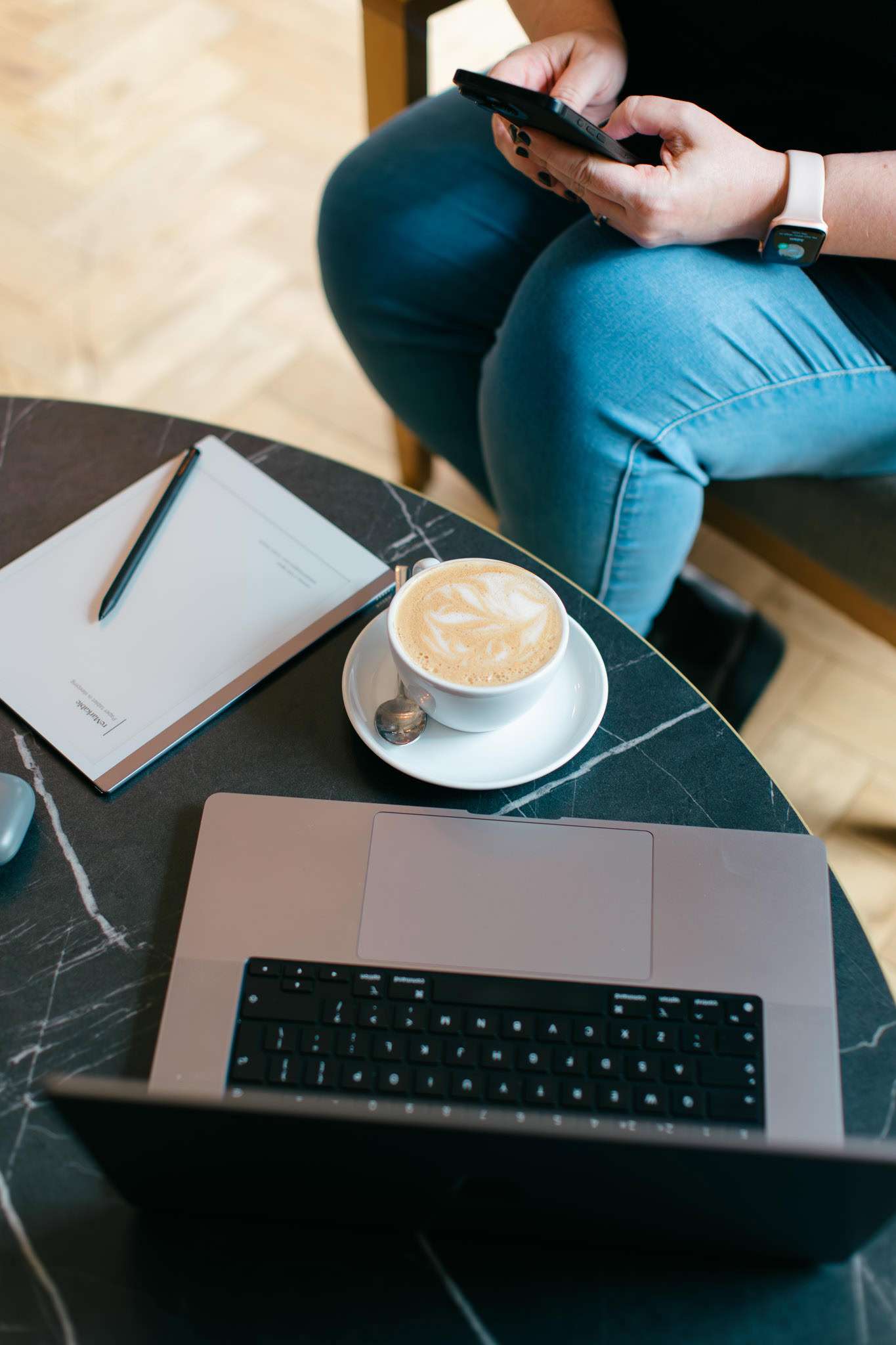 Modern woman working on her mobile phone with a laptop, coffee, and notepad on a black marble table, illustrating professional remote work for service-based businesses.