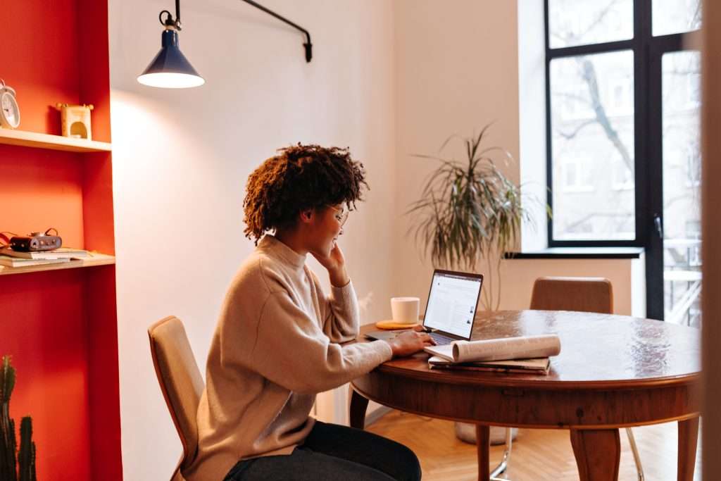 Bright woman working on a laptop at a wooden table in a modern, cosy room with natural light and indoor plants.