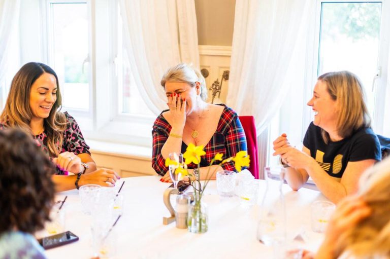 Three women sharing a laugh around a table at a networking event.